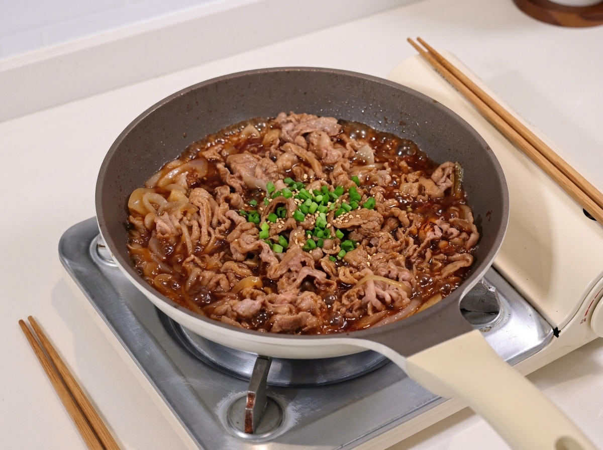 Thinly sliced beef and onions simmering in a dark glossy soy based sauce in a pan.