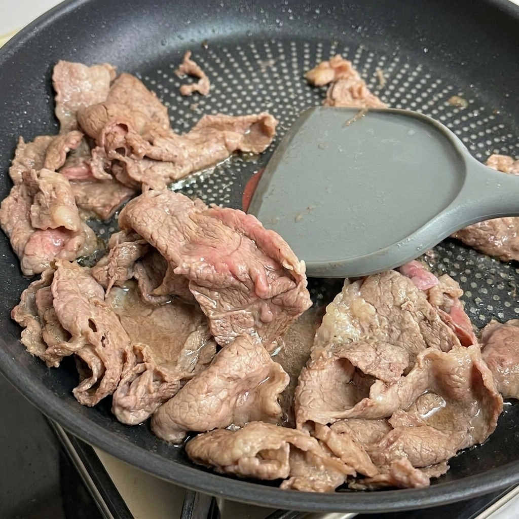 A spatula lifting tender, partially cooked beef strips from the pan.
