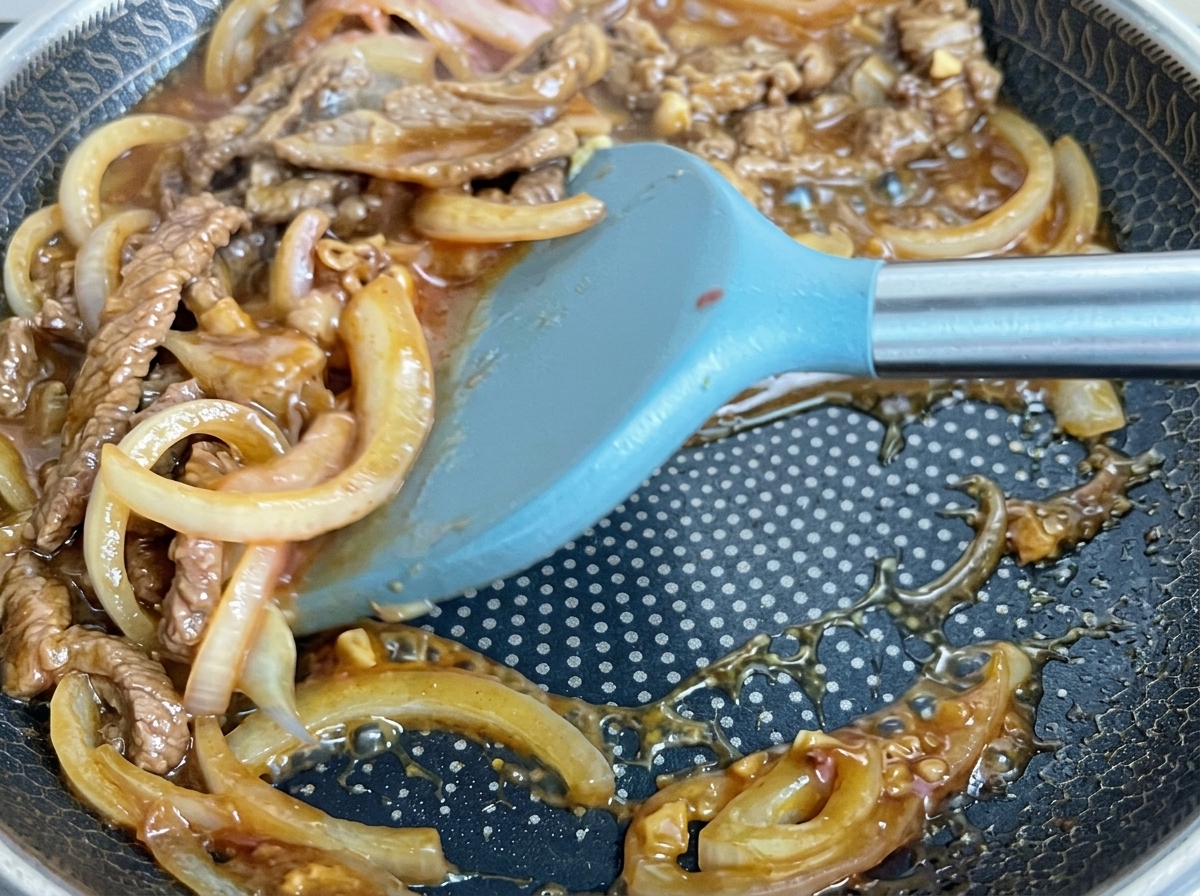 Beef and onions being stirred in a bubbling dark sauce within a pan.