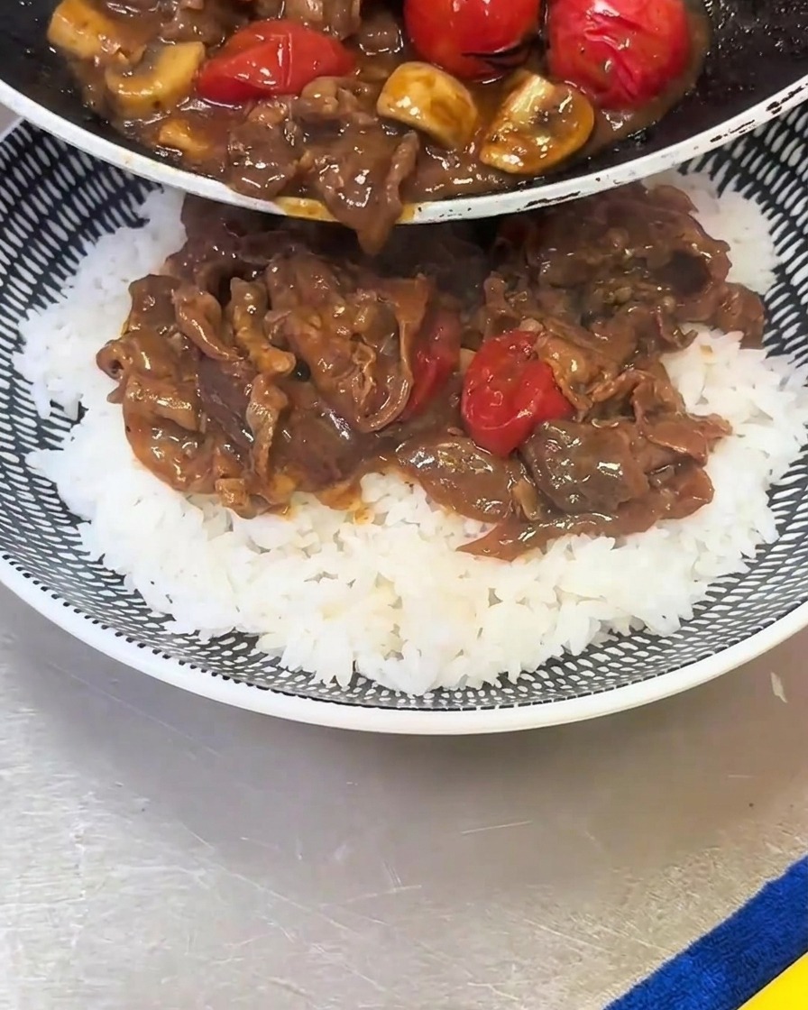 Cooked beef, mushroom, and cherry tomato mixture being poured from a pan onto a bowl of white rice.