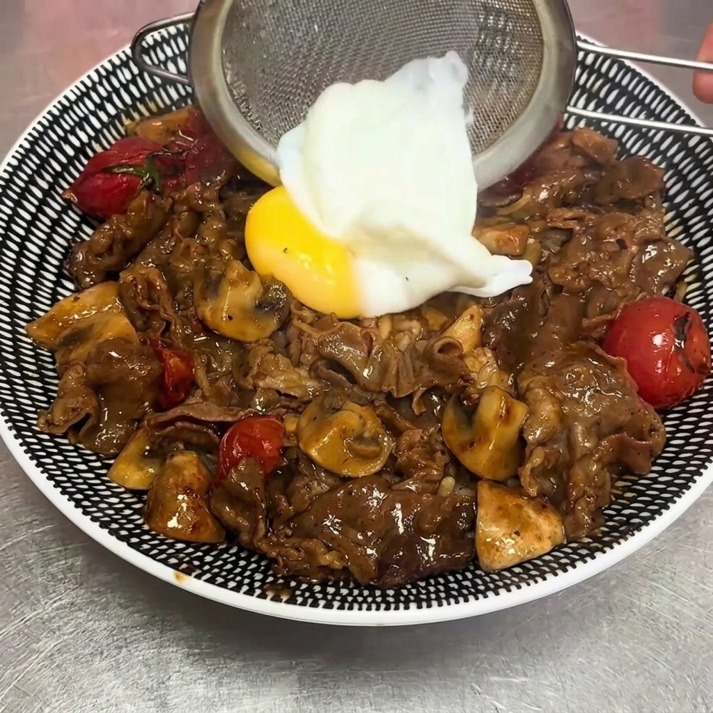 A soft-boiled egg being carefully placed on top of the beef and rice bowl using a wire strainer.