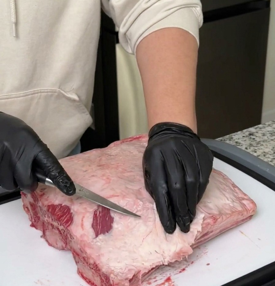 A person wearing black gloves using a sharp boning knife to trim white fat and silver skin from a large rack of raw beef ribs.