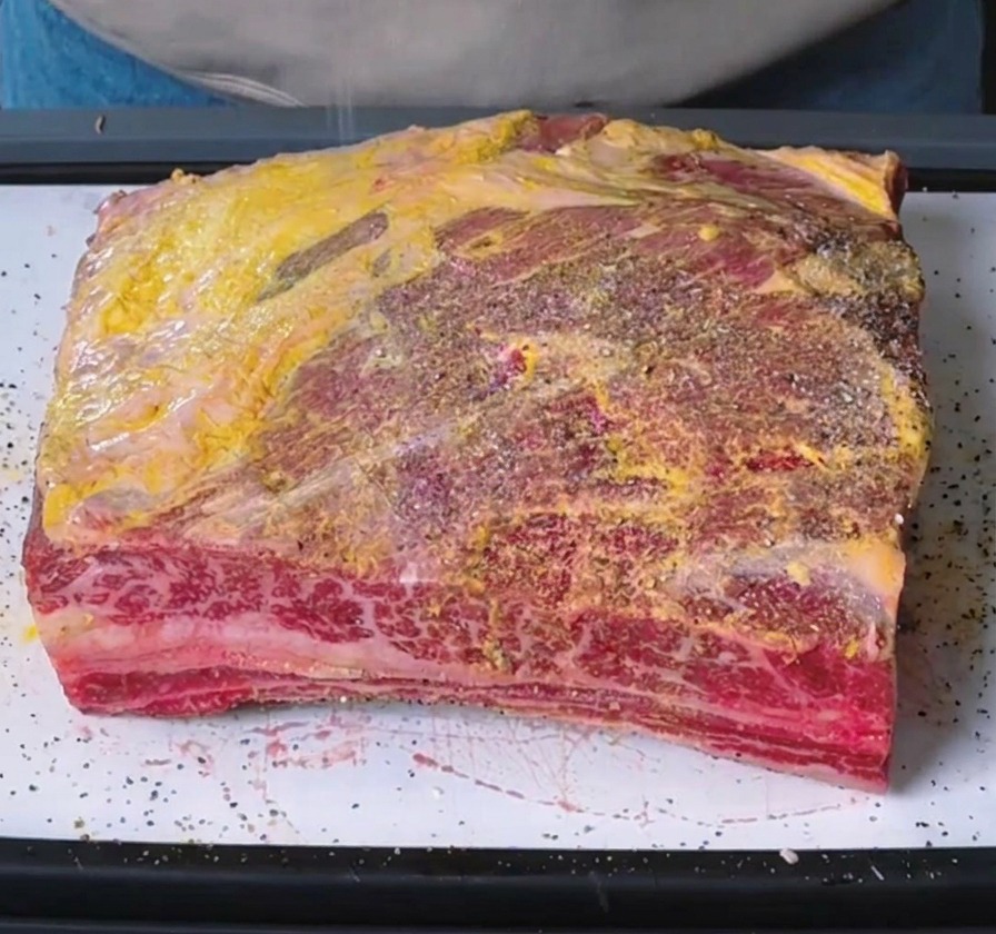 Salt and coarse black pepper being sprinkled onto beef ribs coated with a yellow mustard binder.
