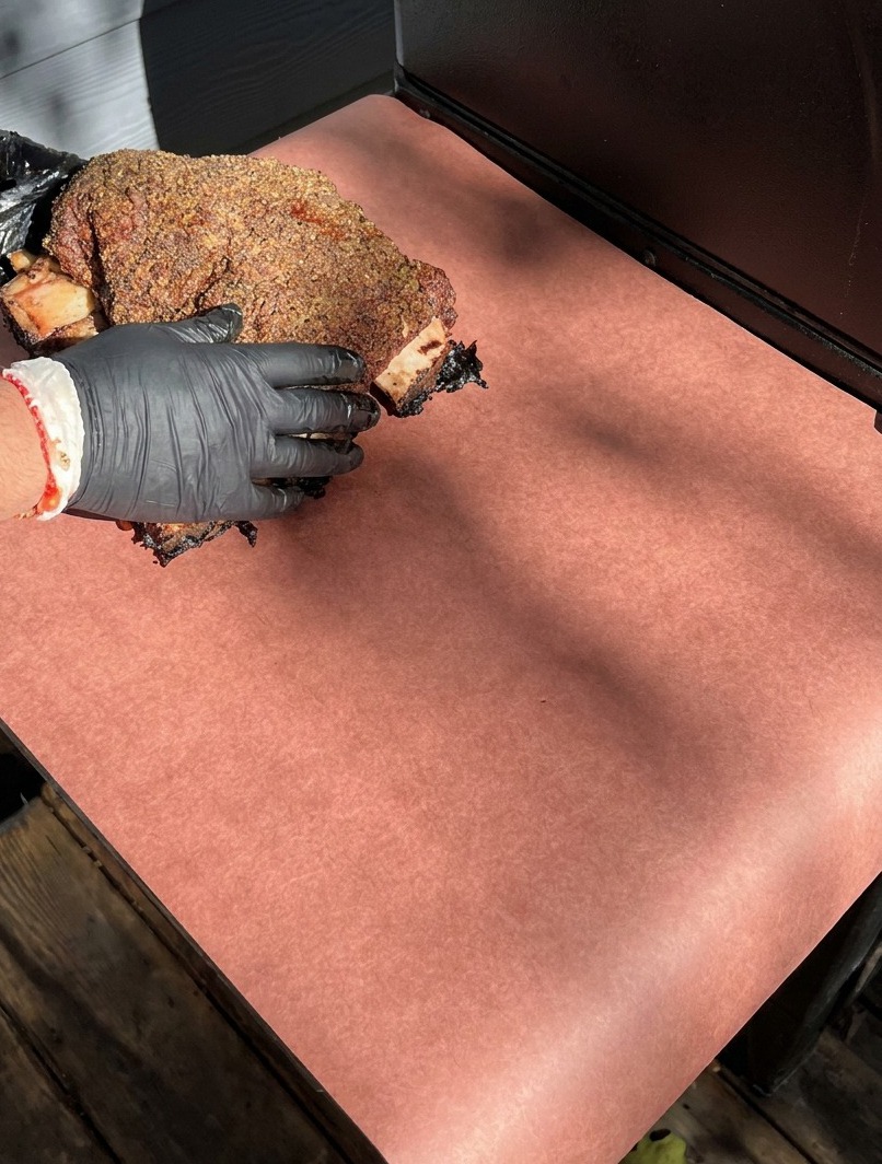 Gloved hands placing dark, smoked beef ribs onto a large sheet of pink butcher paper for wrapping.