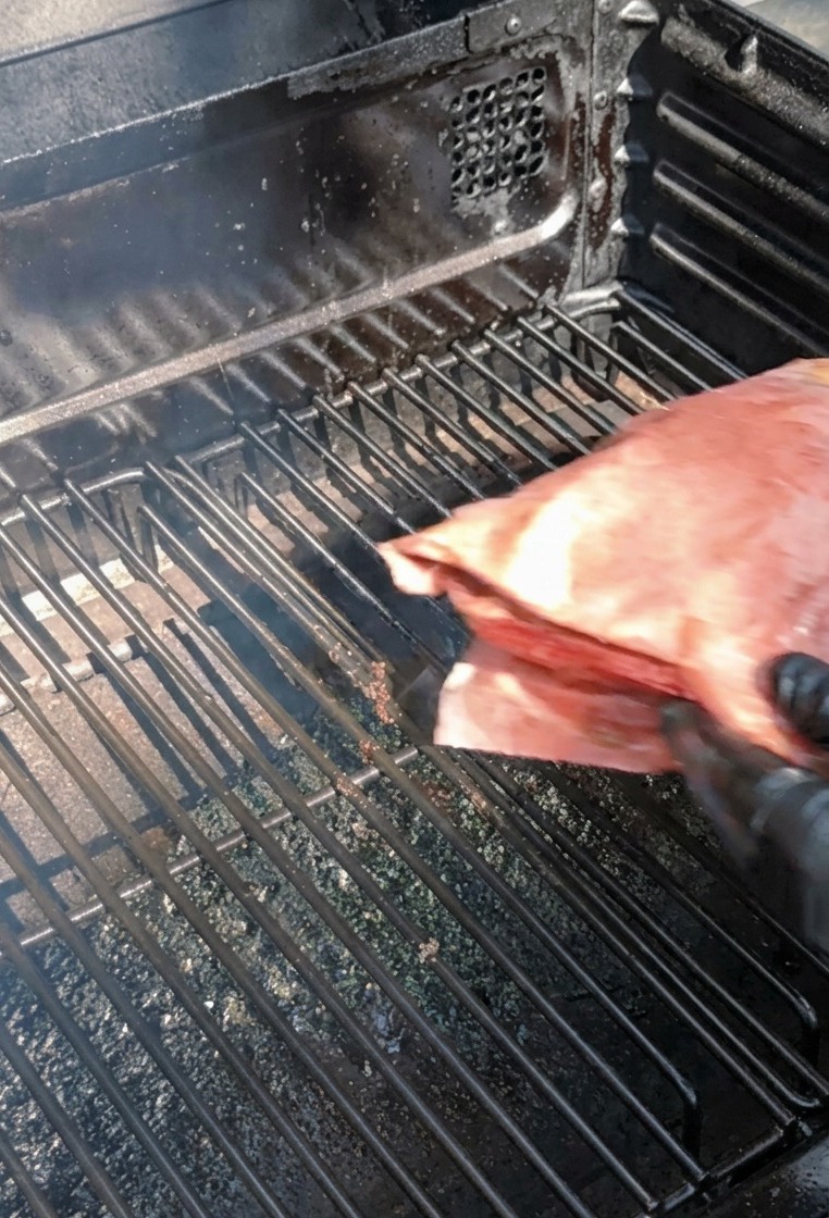 A person wearing black gloves placing a large parcel wrapped in pink butcher paper onto the metal grill grates of a smoker.