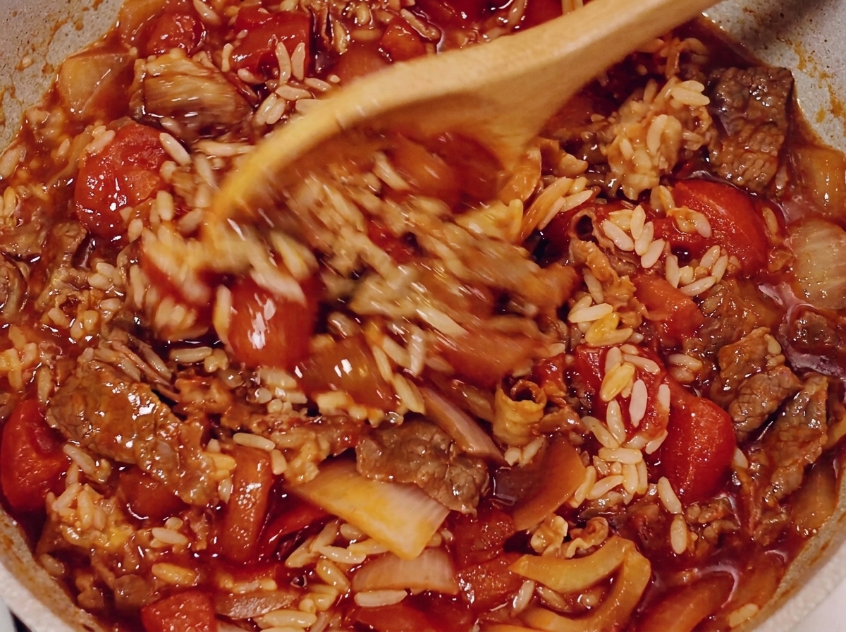 A wooden spoon stirring beef, rice, and tomato sauce in a pan.