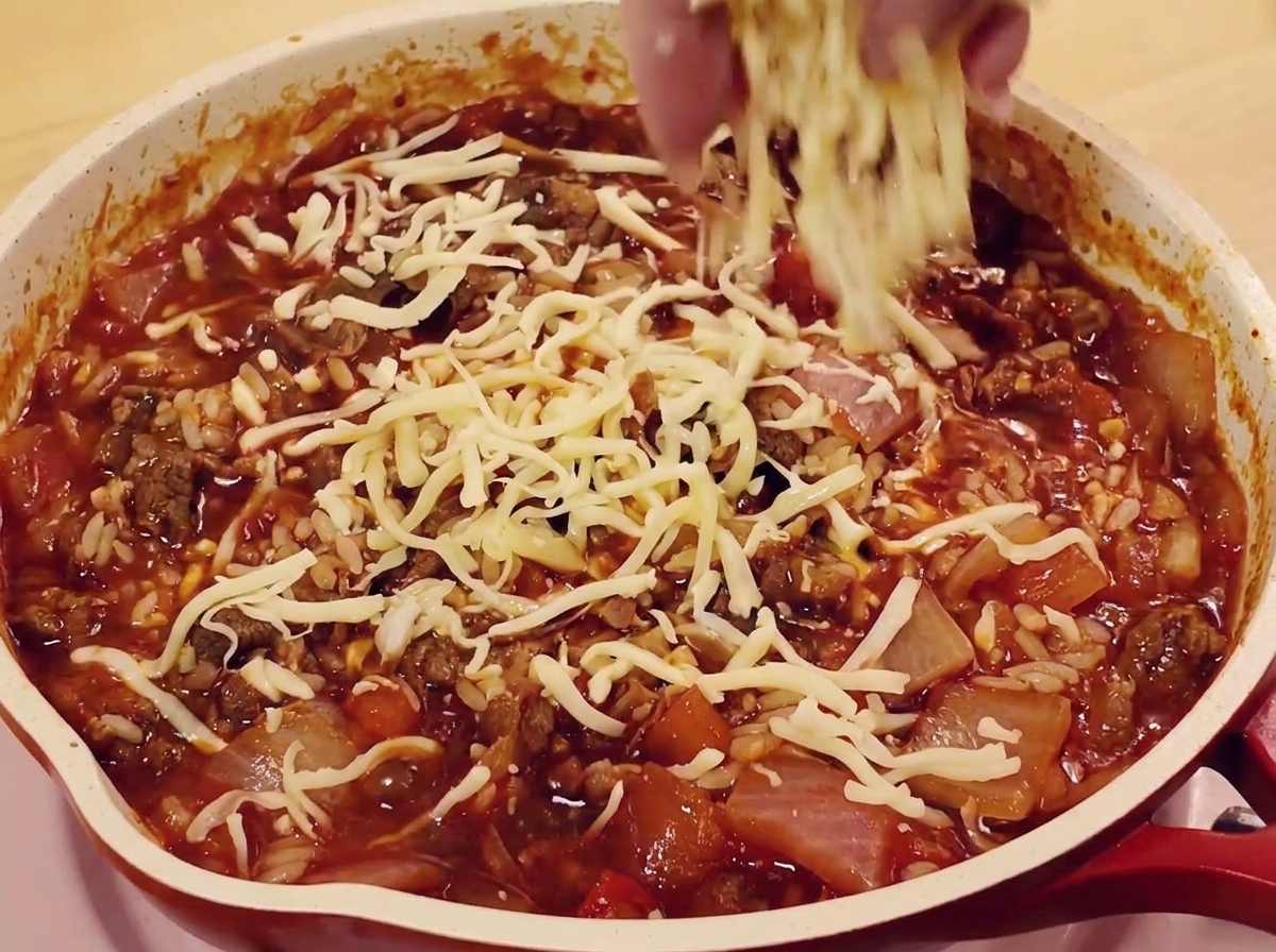 Shredded mozzarella cheese being scattered over a mixture of rice, beef, and tomato sauce in a pan.
