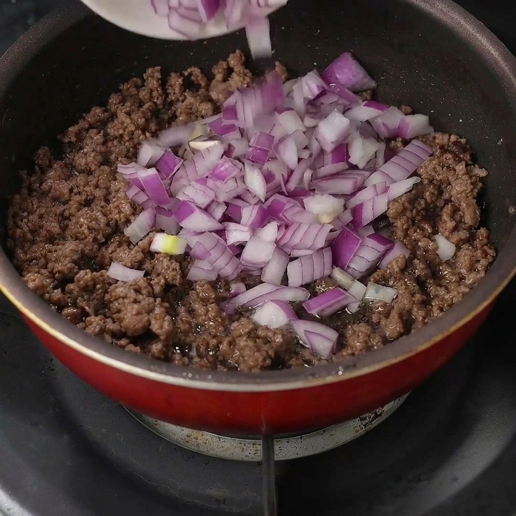 Freshly diced purple onions being poured over cooked ground beef in a frying pan.