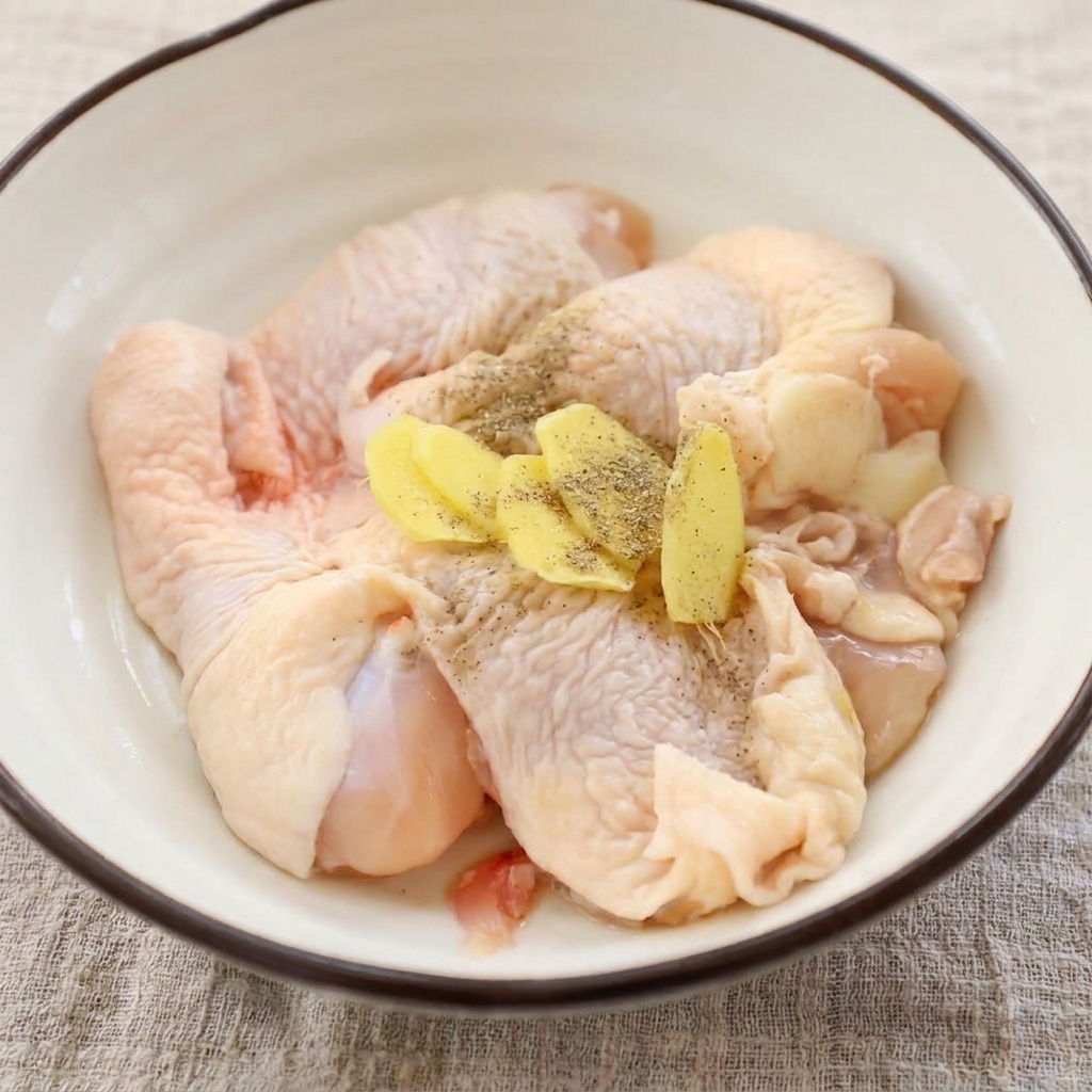 Raw chicken thighs sitting in a bowl, topped with sliced fresh ginger and ground black pepper.