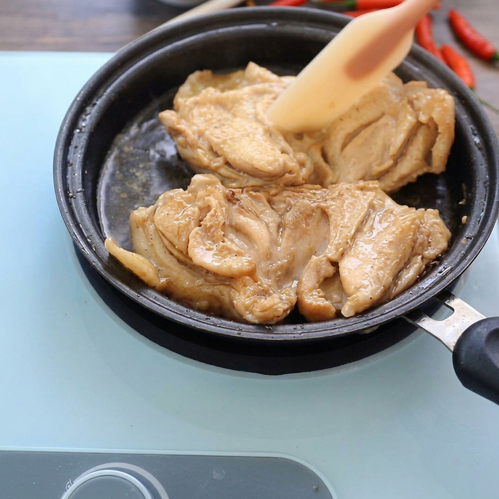 Cooked chicken thighs simmering in a shallow pan with dark sauce, being lightly pressed with a silicone spatula.