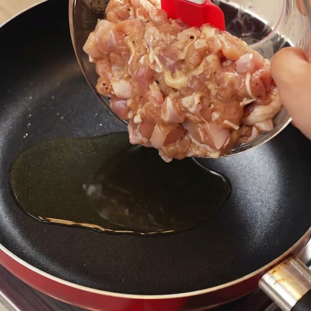 Using a red spatula to transfer marinated raw chicken pieces from a glass bowl into a hot frying pan with oil.