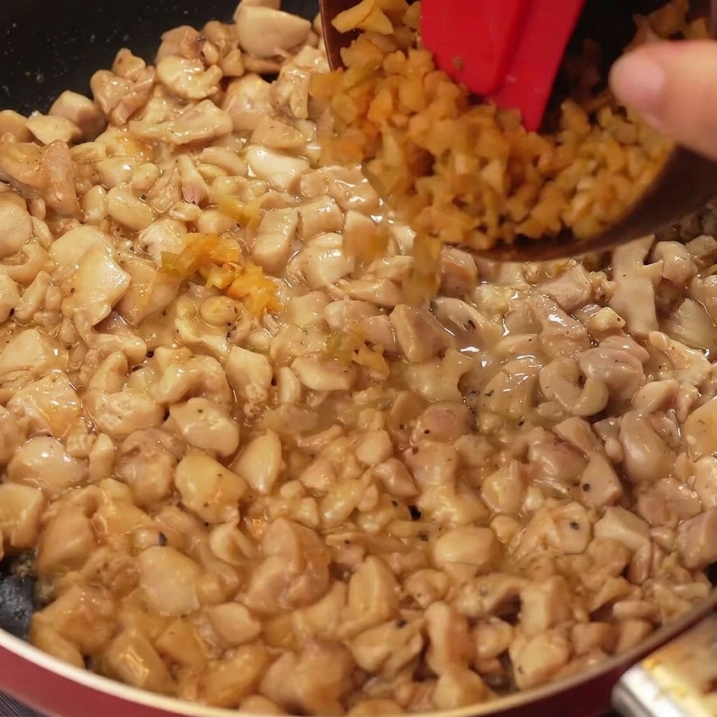 Finely chopped preserved mustard greens and white onion pieces being added to a pan of cooked, golden-brown chicken pieces.
