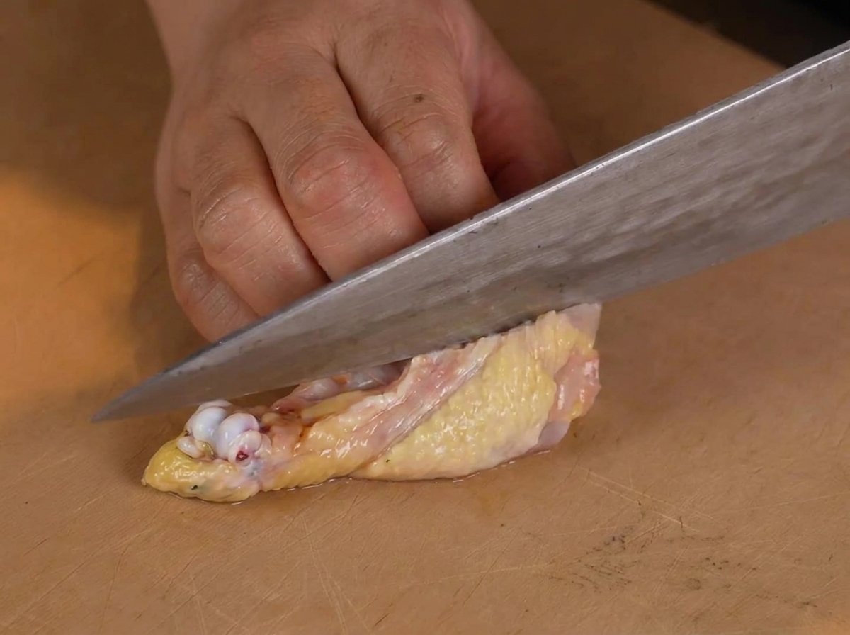 A person using a chefs knife to carefully remove the bone from a raw chicken leg on a wooden cutting board.