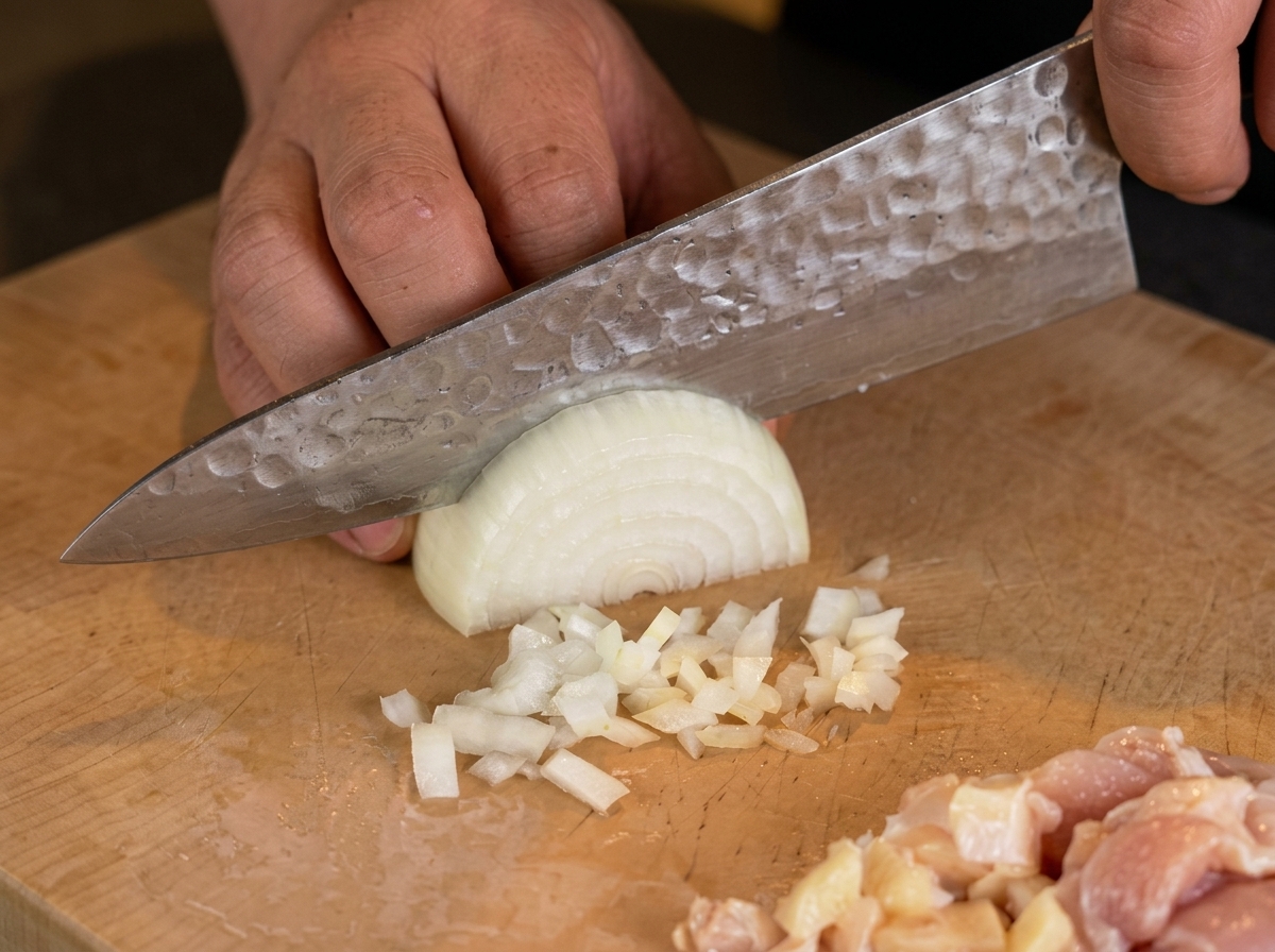 A chefs hand using a knife to finely chop a half of a white onion on a wooden board.