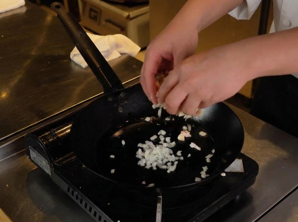 Two hands adding prepared diced raw chicken and cartilage into the hot pan with sautéing onions.