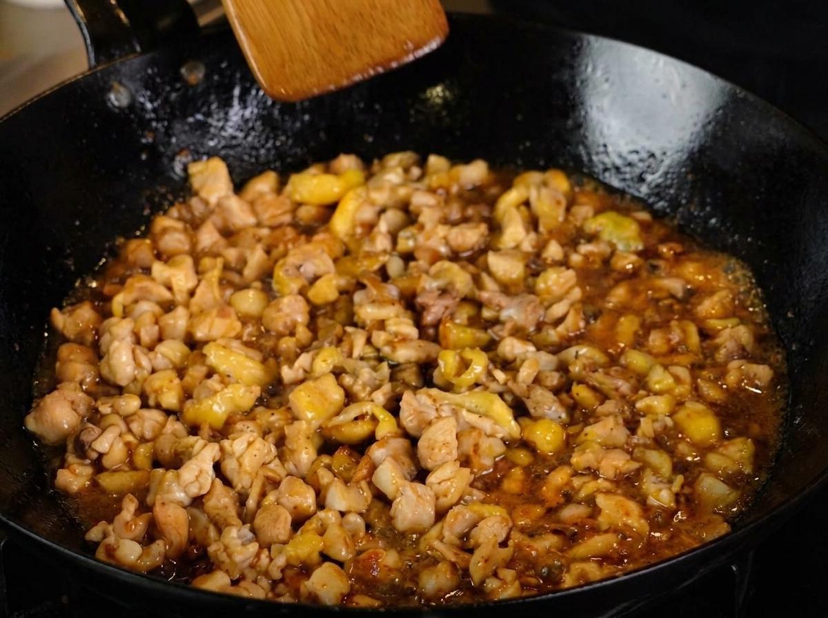 Diced chicken simmering in a rich dark sauce, sprinkled with black pepper, being stirred with a wooden spatula.