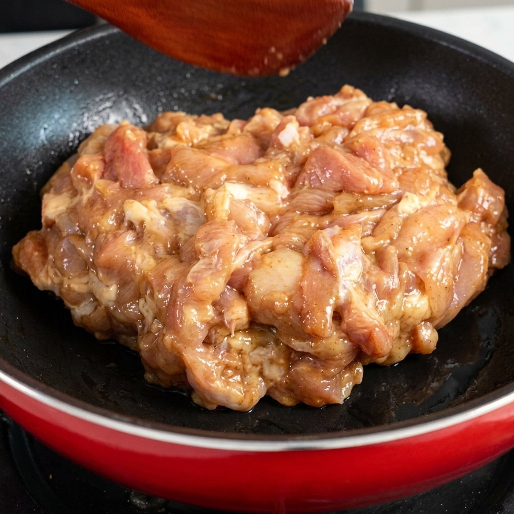 Marinated raw chicken pieces being placed into a heated red frying pan.