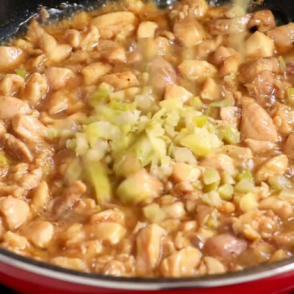 Finely diced pickled mustard greens being added to a pan with partially cooked chicken pieces.
