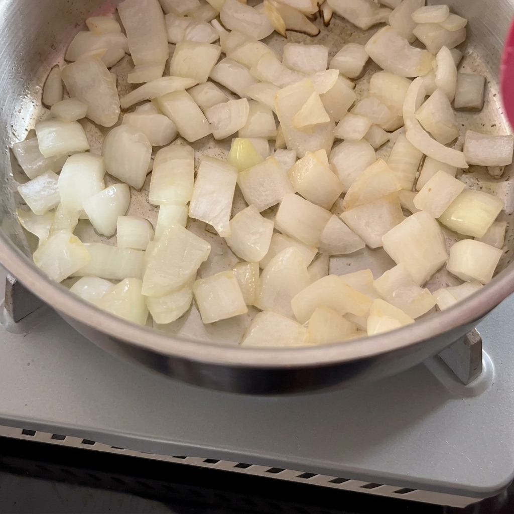Chopped white onions being sautéed in a metal pot with a red spatula.