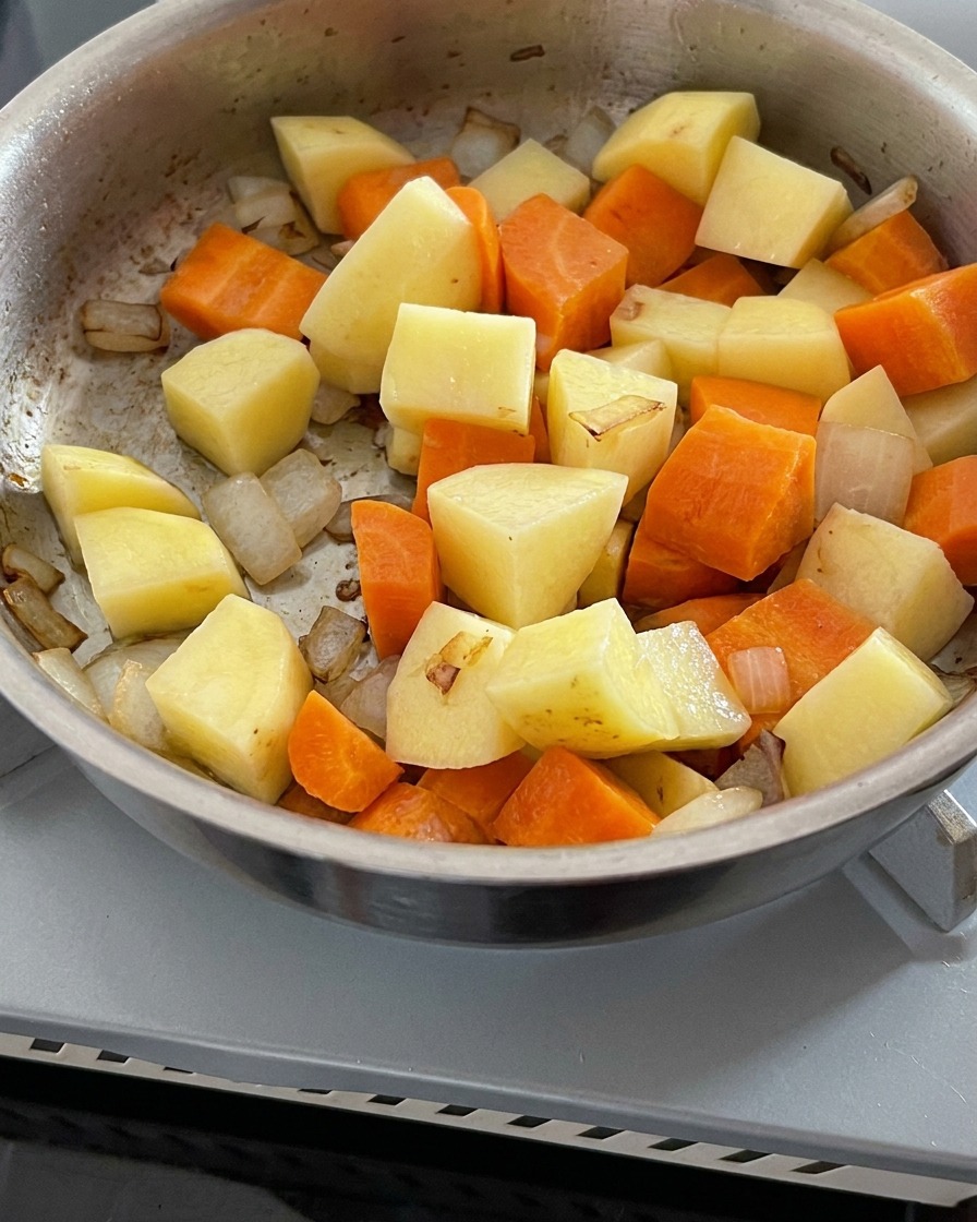 Clear water being poured into a pan containing sautéed onions, carrots, and potatoes.