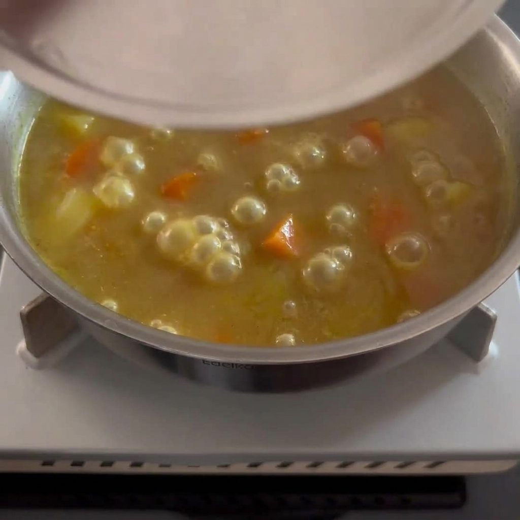 A thick, golden-brown curry sauce being stirred with pieces of soft potato and carrot visible.