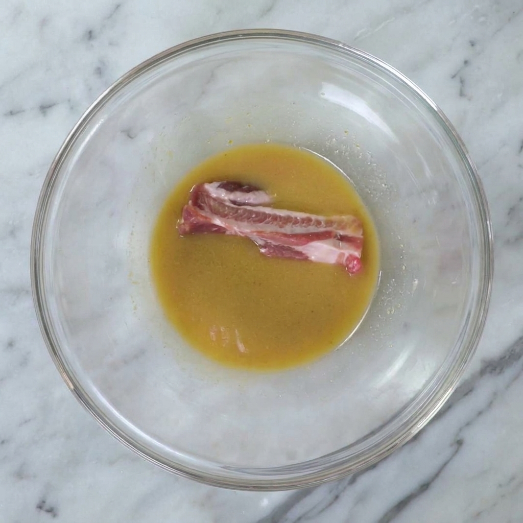 A single raw pork rib being submerged into the smooth golden miso marinade inside a large glass bowl