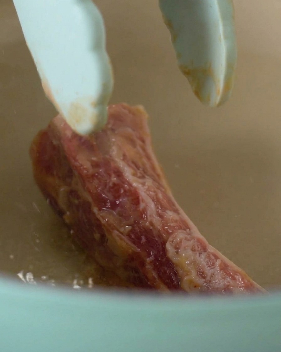 Tongs placing a single piece of marinated raw pork rib into a heated light blue pot.