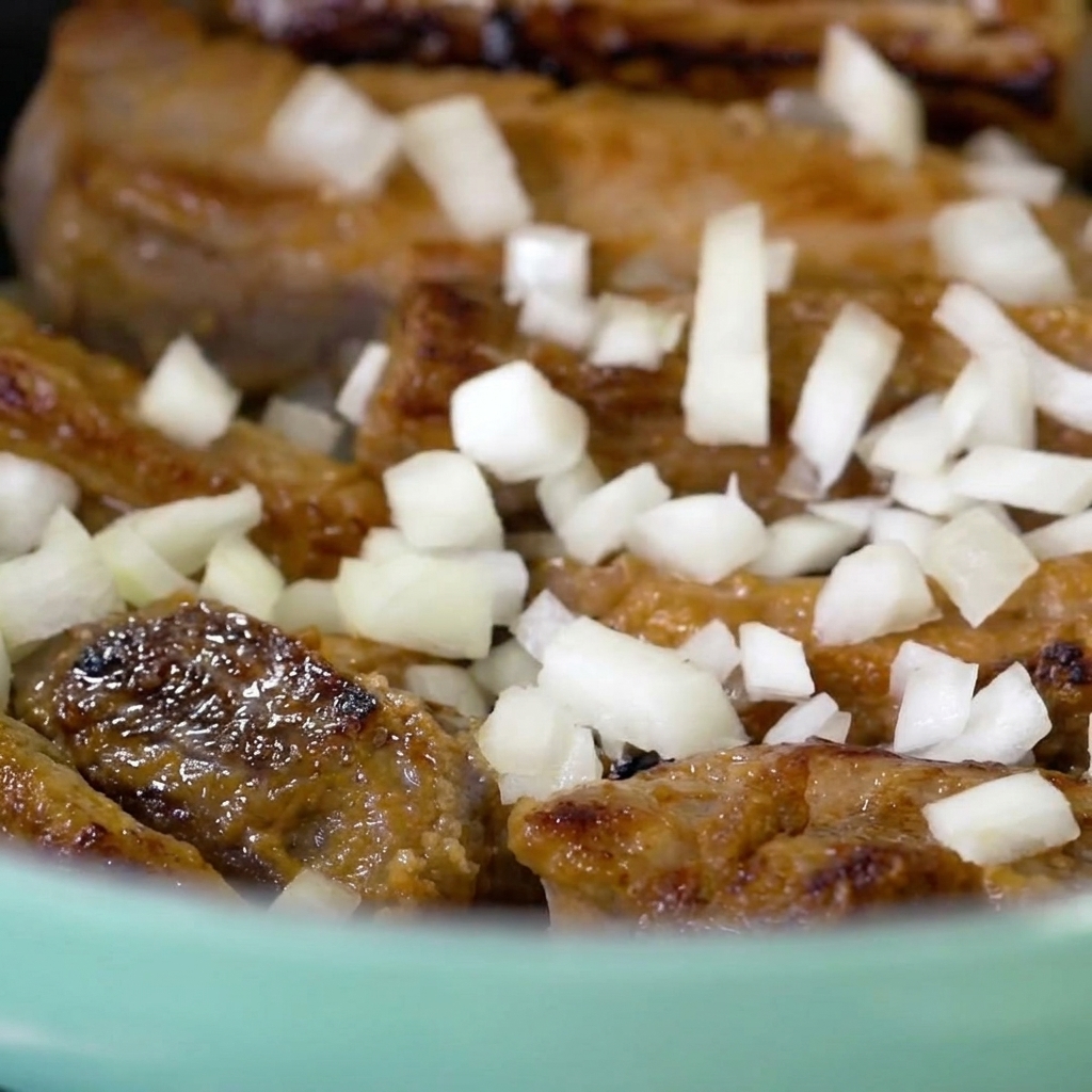 Freshly chopped white onion pieces being scattered over seared pork ribs inside a light blue pot.