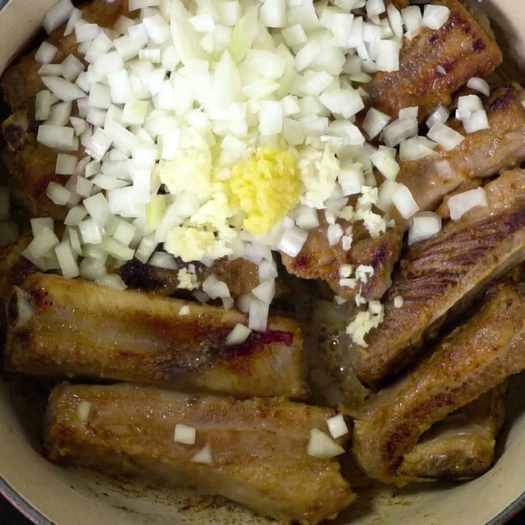 Top-down view of pork ribs in a pot, generously covered with diced white onions and a mound of minced yellow ginger and garlic.