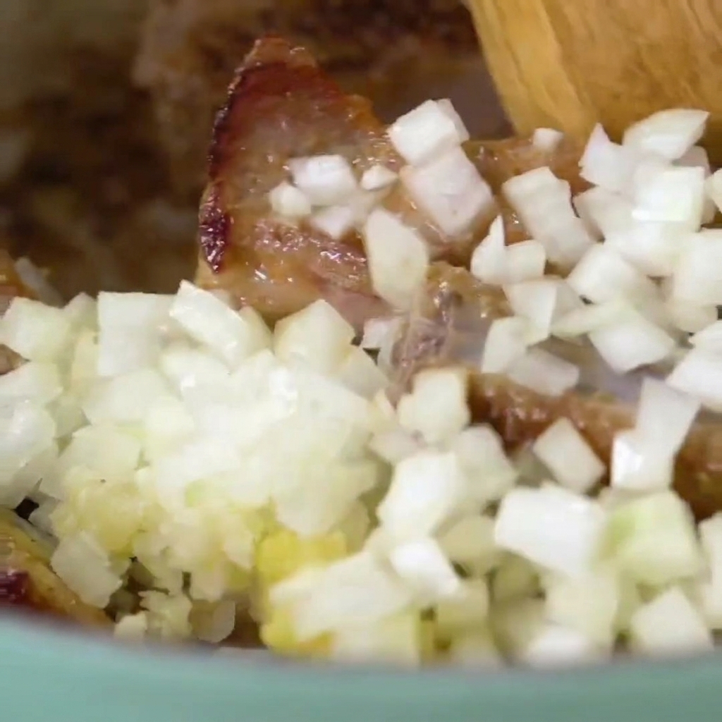 Close-up of a wooden spoon tossing browned pork ribs with chopped onions and minced garlic in a cooking pot.