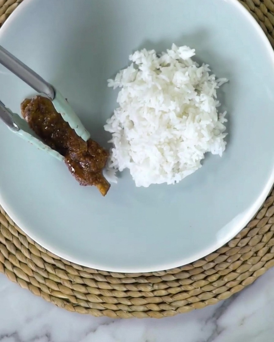 Silicone-tipped tongs placing a sticky, dark glazed pork rib onto a light blue plate next to fluffy white rice.