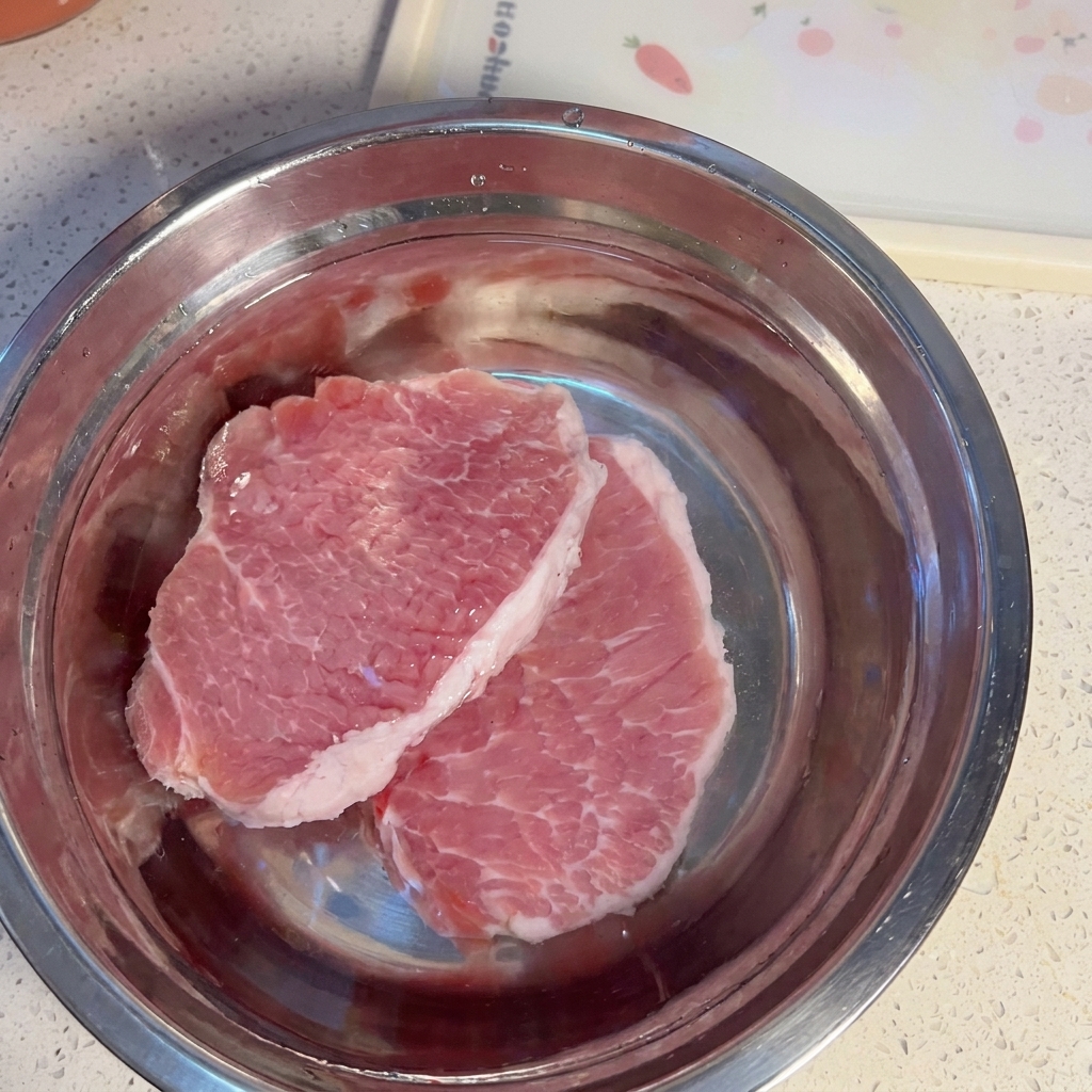 Raw pork slices soaking in a metal bowl filled with water.