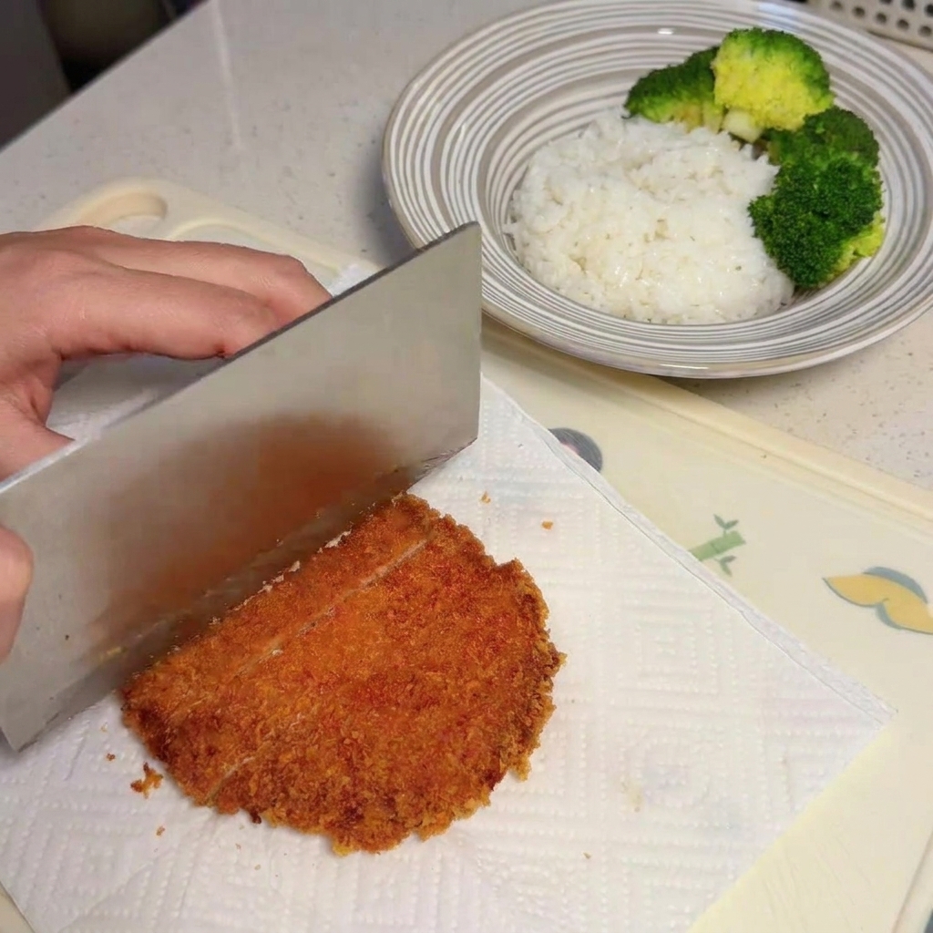 A hand using a large knife to cleanly slice a crispy golden-brown fried pork chop into strips on a paper towel.