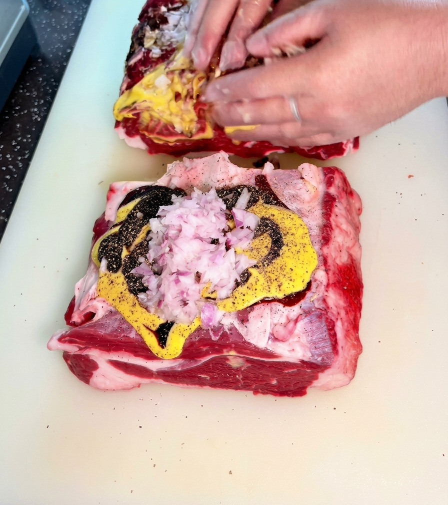 Hands actively rubbing a mixture of chopped onions, mustard, and dark sauces into large slabs of raw beef.