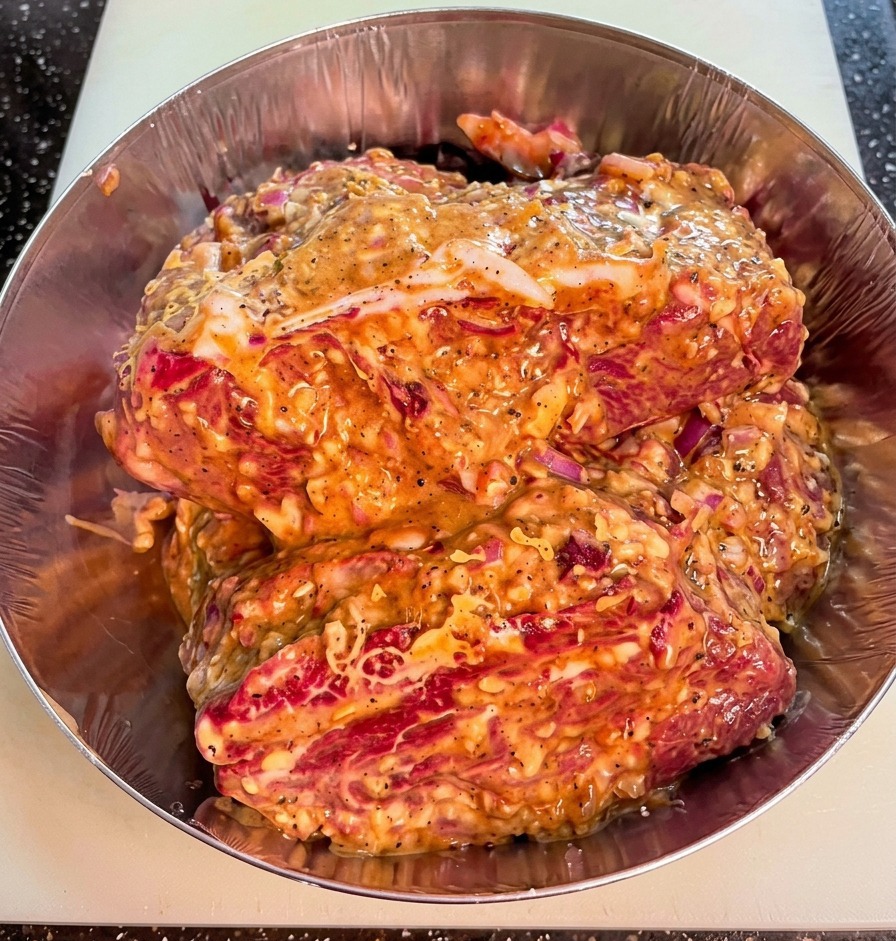 A close-up shot of raw beef chuck pieces in a metal bowl, coated in a thick, textured marinade with visible bits of red onion and cracked black pepper.