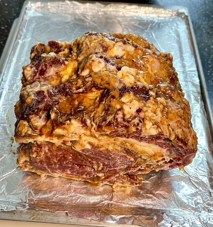 A thick slab of marinated beef chuck coated in sauce and onions, sitting on a tray lined with crinkled aluminum foil.