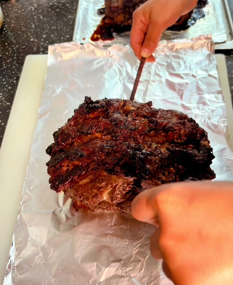 A metal skewer piercing the dark, charred surface of a large beef roast resting on aluminum foil.