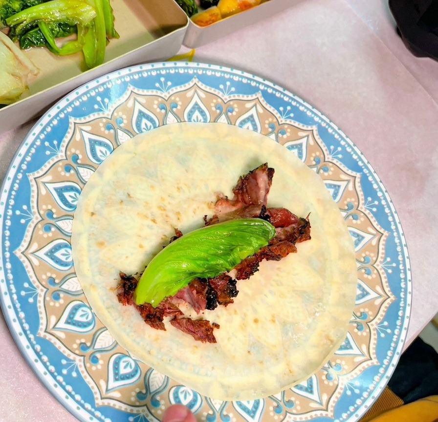 A top-down view of a blue patterned plate with a tortilla topped with juicy slices of smoked beef and a fresh green leaf.
