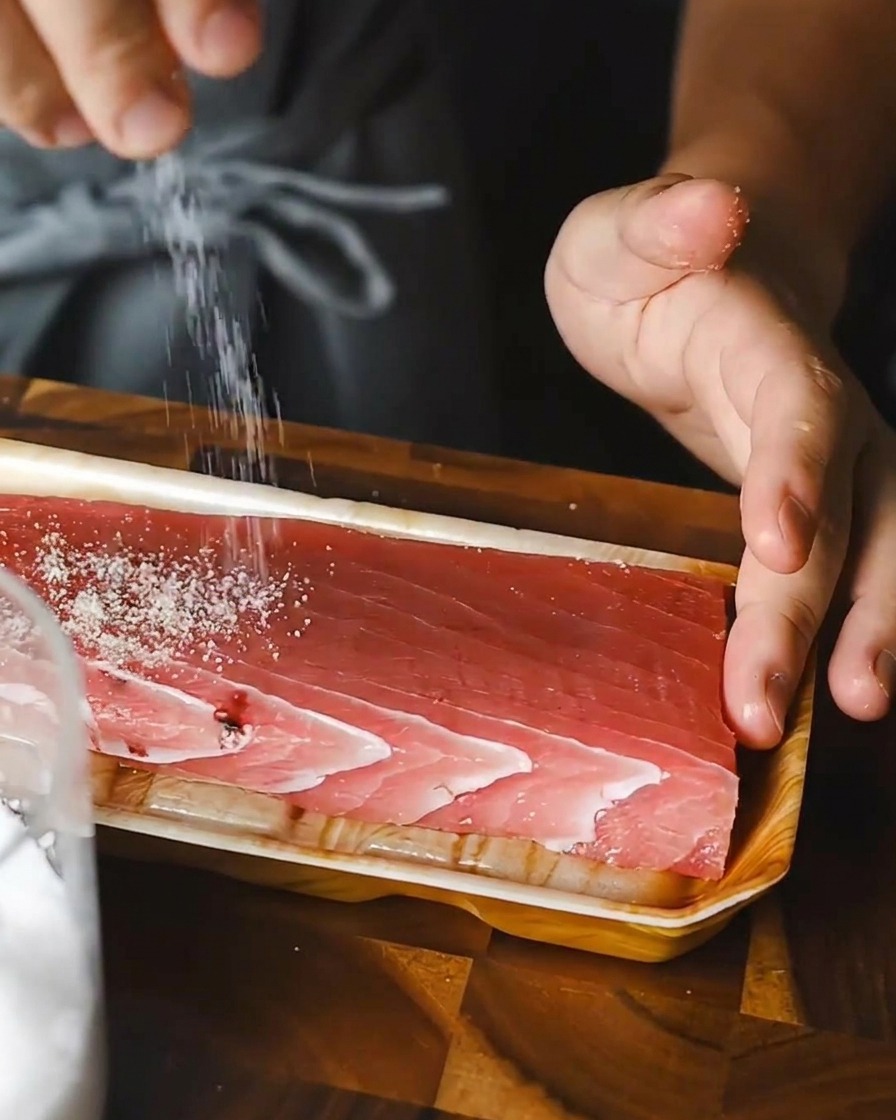 A person sprinkling salt evenly over a rectangular block of raw red tuna on a tray.