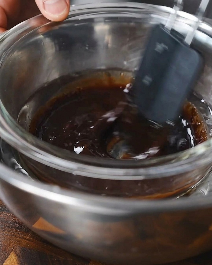 A spatula stirring a dark liquid marinade in a glass bowl set over a water bath.