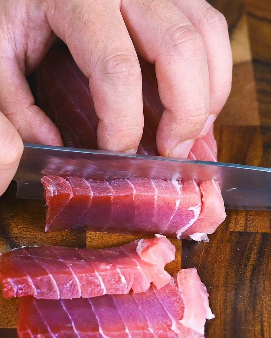 Hands using a chefs knife to slice a block of raw red tuna into thick, even rectangles on a wooden cutting board.