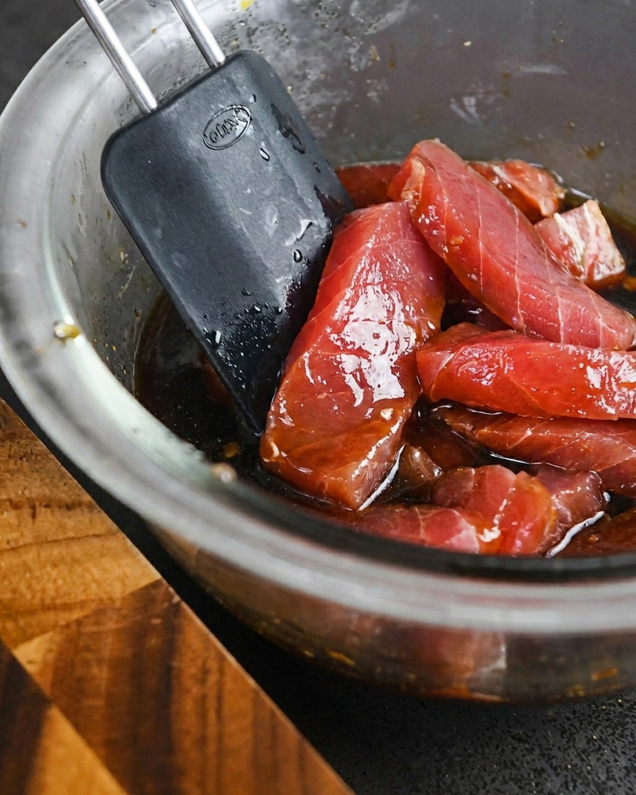 Thick slices of raw red tuna being mixed into a bowl of dark liquid marinade with a spatula.