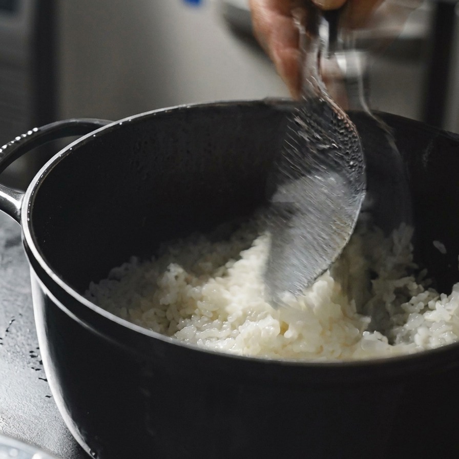 A hand using a clear rice paddle to stir and scoop steaming hot white rice inside a black cooking pot.