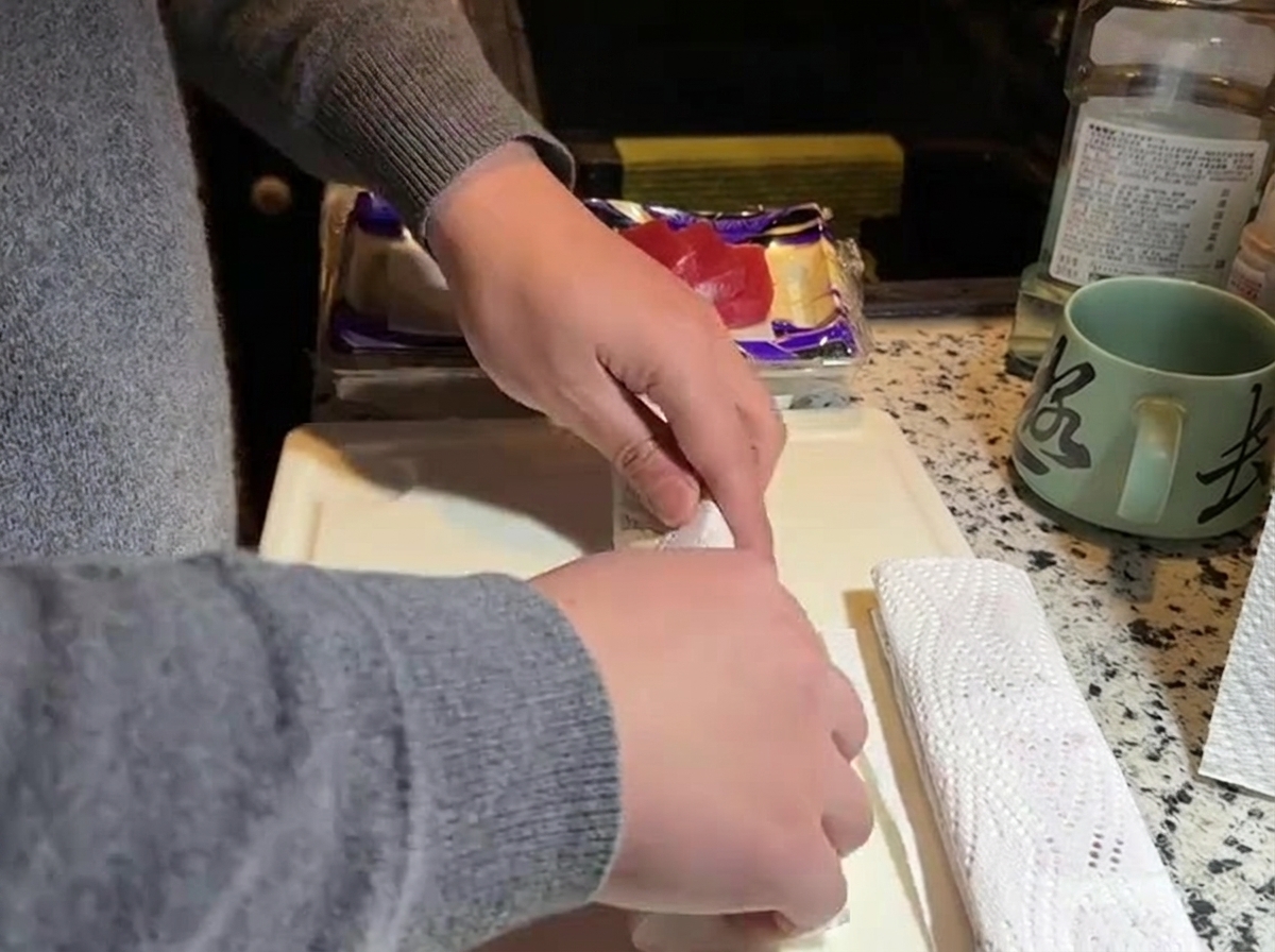 Hands pressing white kitchen paper towels against a block of raw red tuna on a white cutting board.