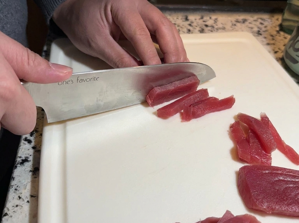 A sharp chefs knife slicing a piece of bright raw red tuna into long strips on a white cutting board.
