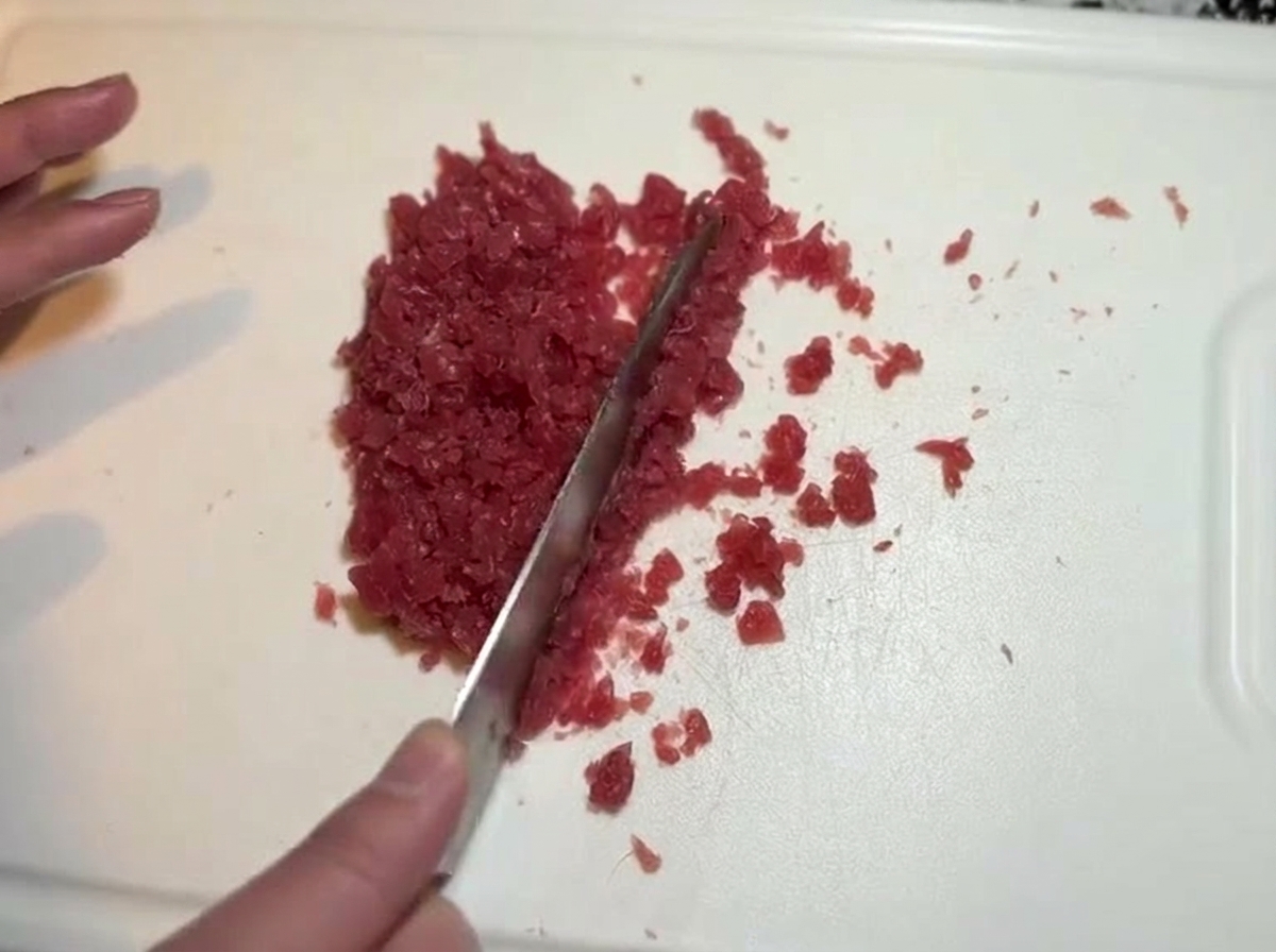 A knife finely dicing and chopping pieces of raw red tuna into a fine mince on a cutting board.