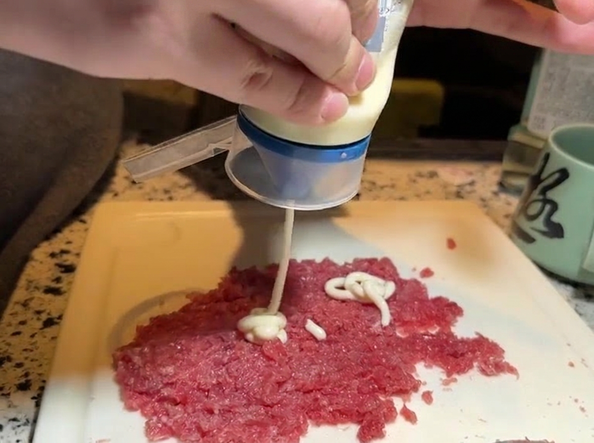 Squeezing white mayonnaise from a bottle onto a separated portion of freshly minced raw red tuna.