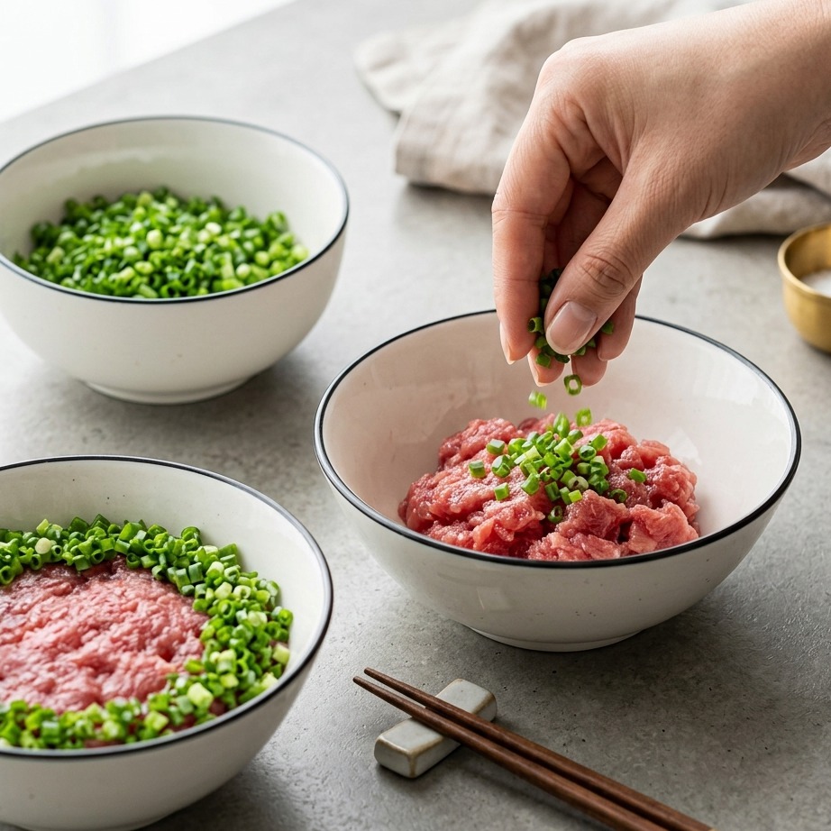 A close-up view of fresh green scallions being sprinkled onto a bowl of minced tuna.