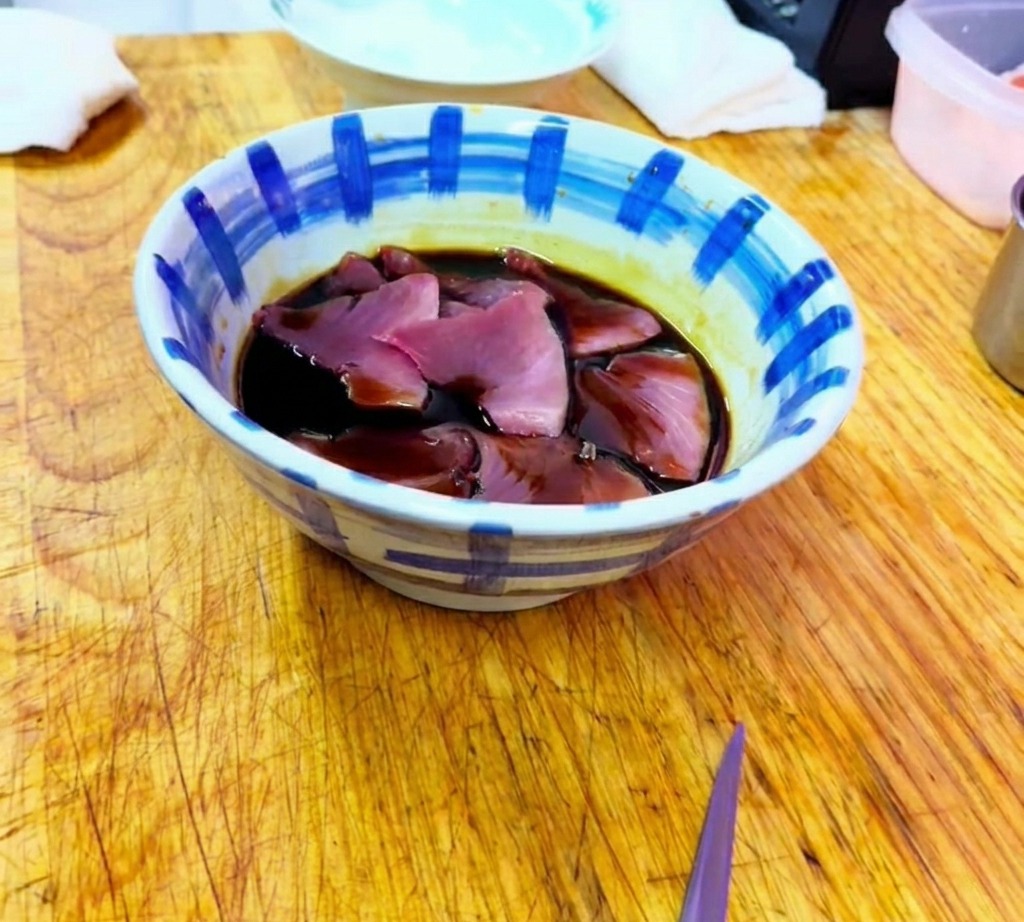 Raw tuna slices being submerged in a dark, savory soy-based marinade within a patterned ceramic bowl.
