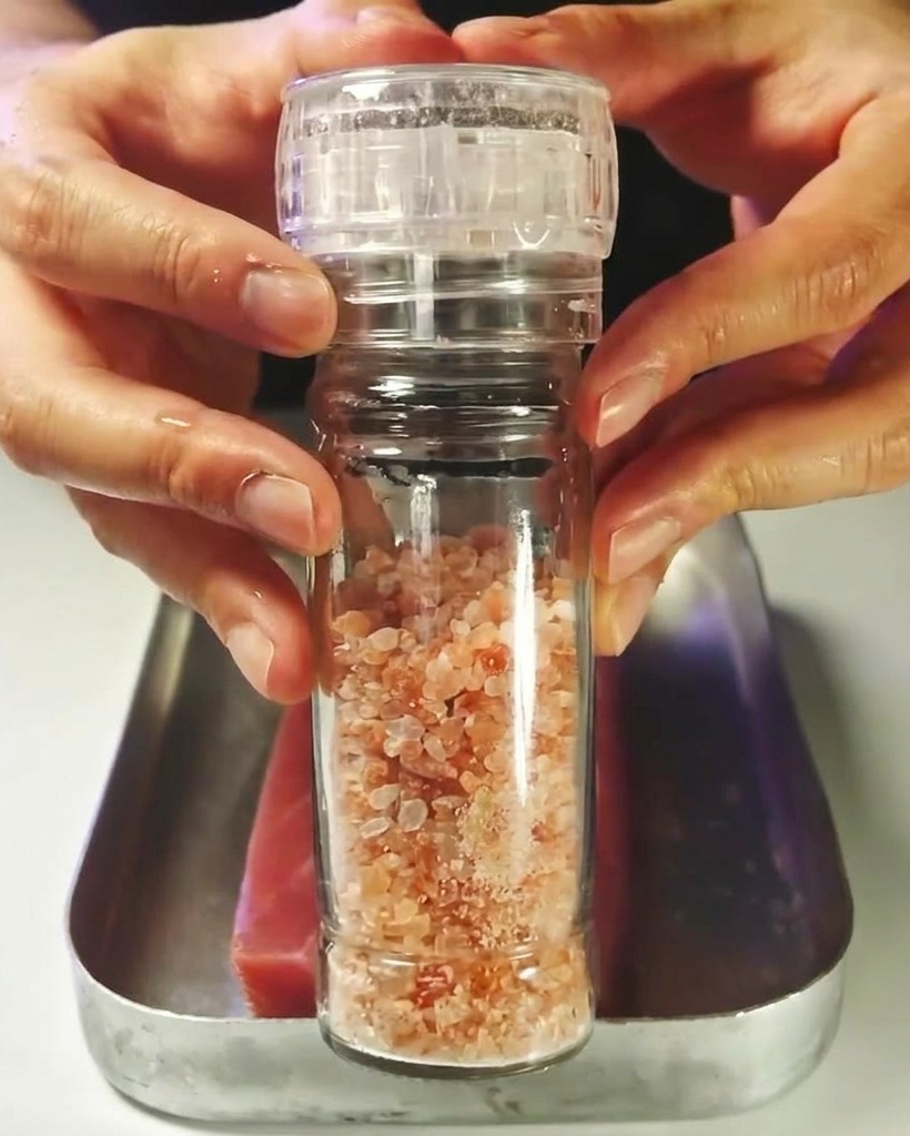A block of raw red tuna being seasoned with salt from a mill.